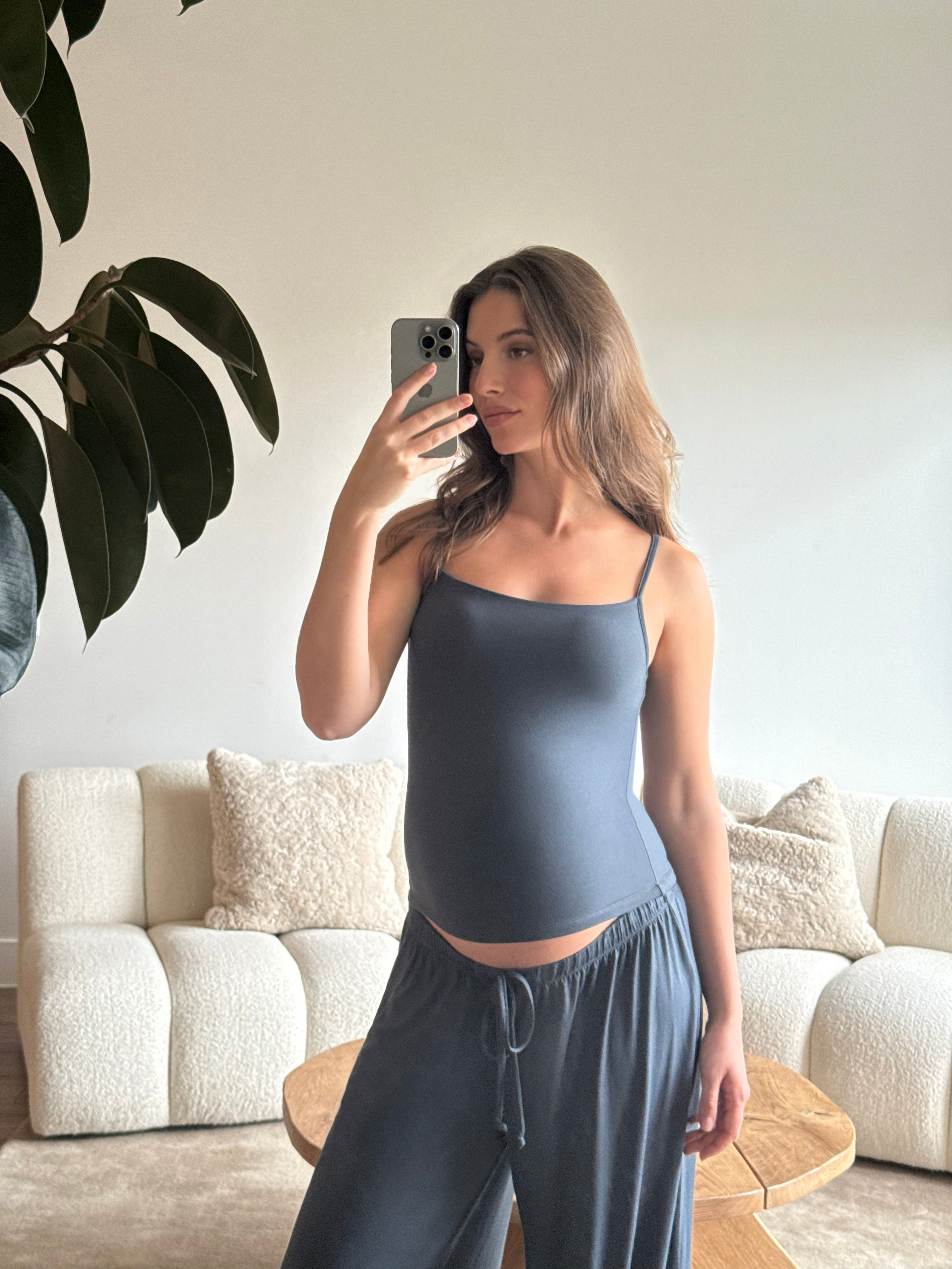 Pregnant woman taking a mirror selfie wearing a Bumpsuit cloud cement blue tank top and pants in a living room.