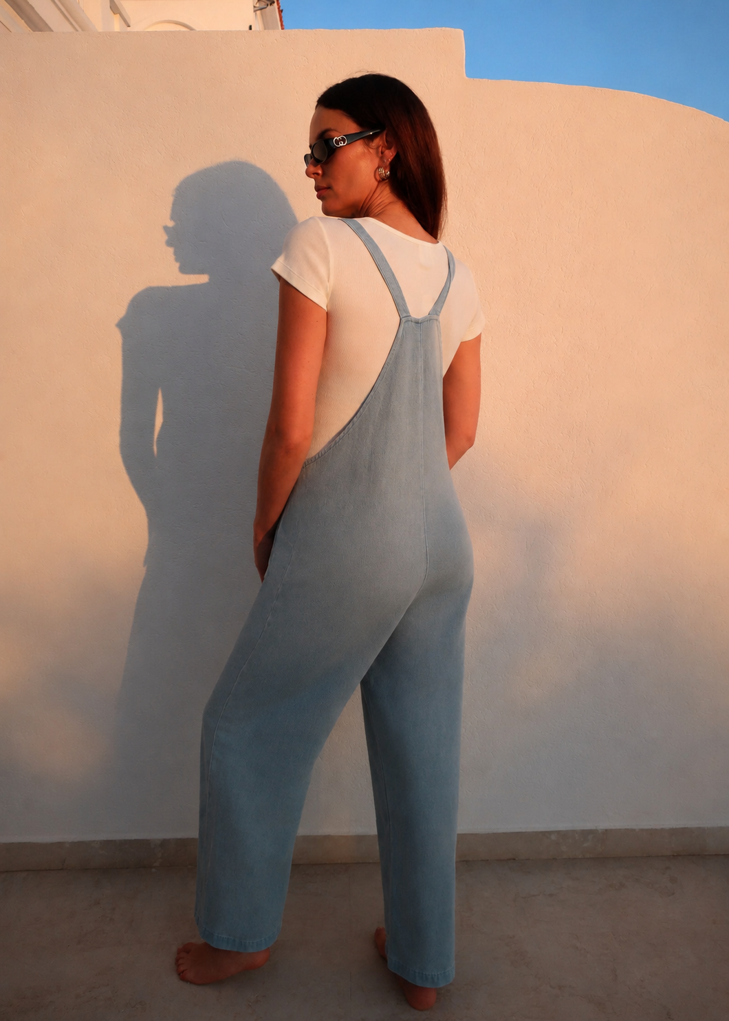 A woman stands barefoot against a white wall, wearing a Bumpsuit light blue denim overall jumpsuit over a white t-shirt and sunglasses. The warm sunlight casts a long shadow of the model onto the wall.