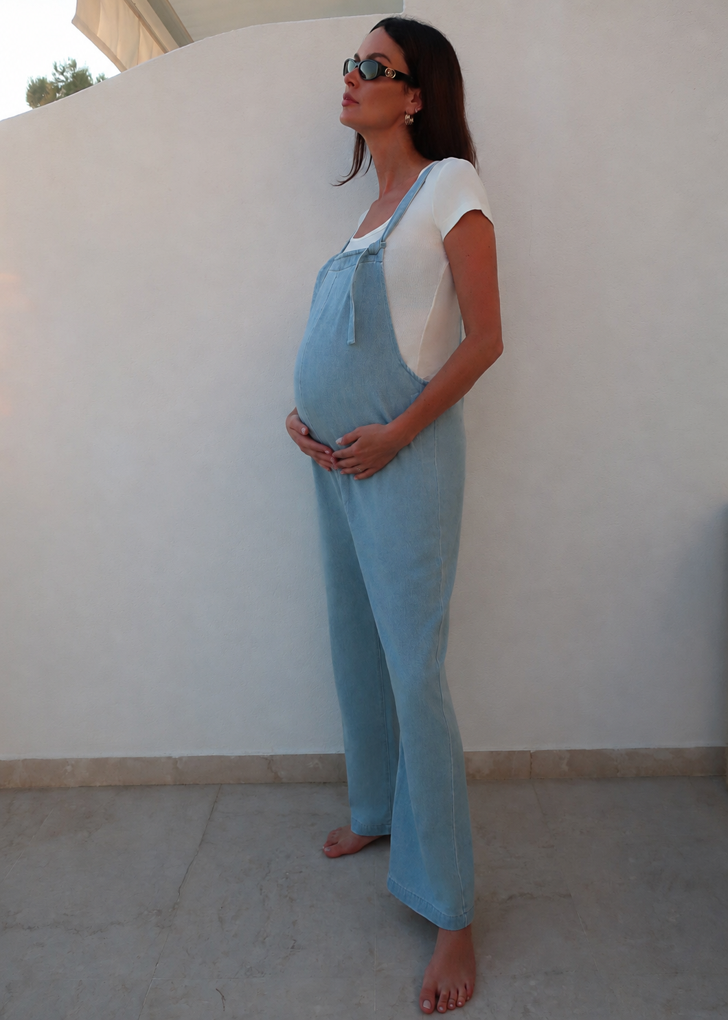 A pregnant woman wearing a light blue linen overall jumpsuit over a white t-shirt by Bumpsuit, standing barefoot against a white stucco wall.
