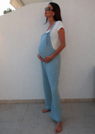 A pregnant woman wearing a light blue linen overall jumpsuit over a white t-shirt by Bumpsuit, standing barefoot against a white stucco wall.