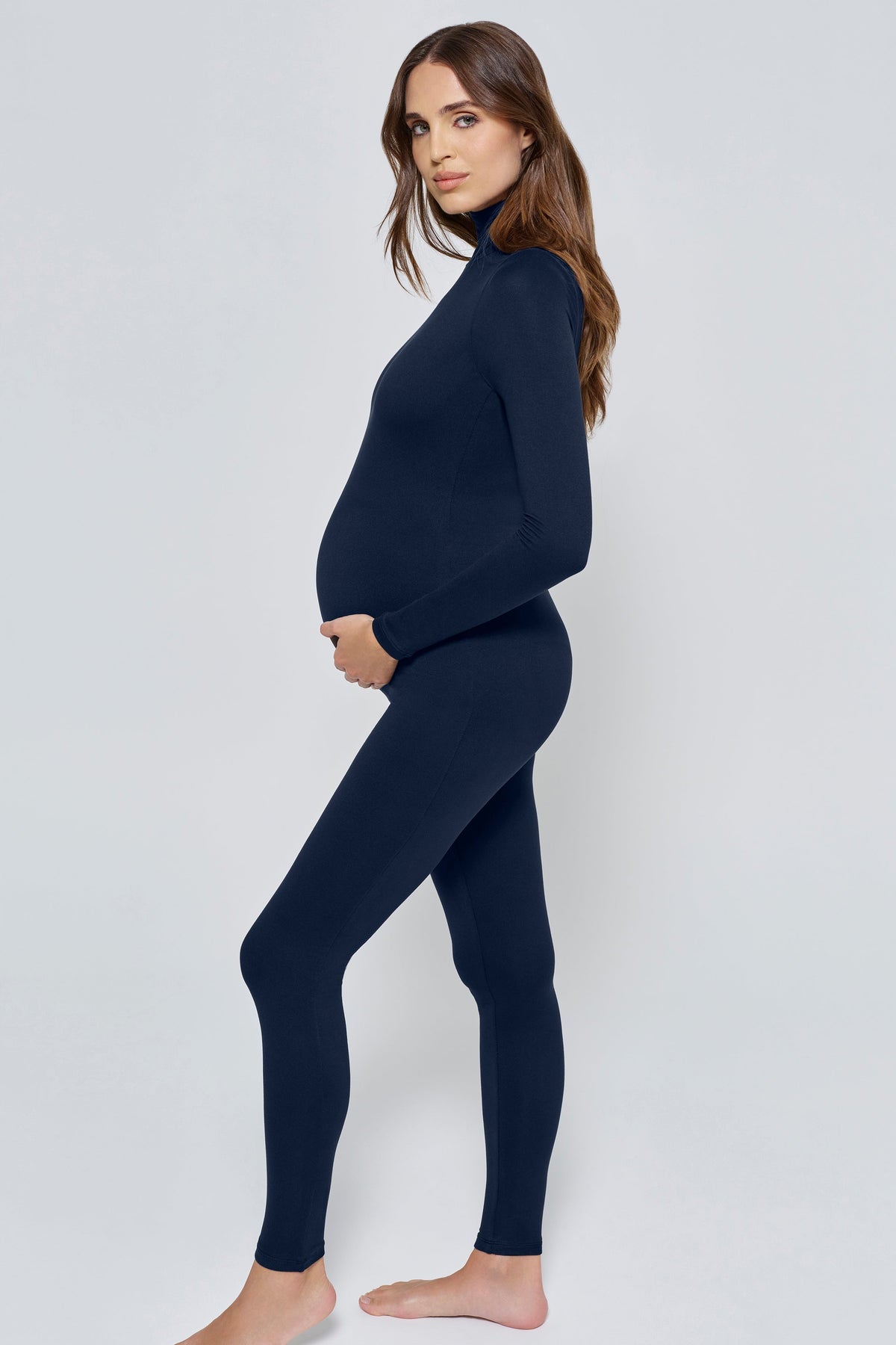 A pregnant woman modeling a dark navy blue maternity turtleneck jumpsuit against a light gray studio background.