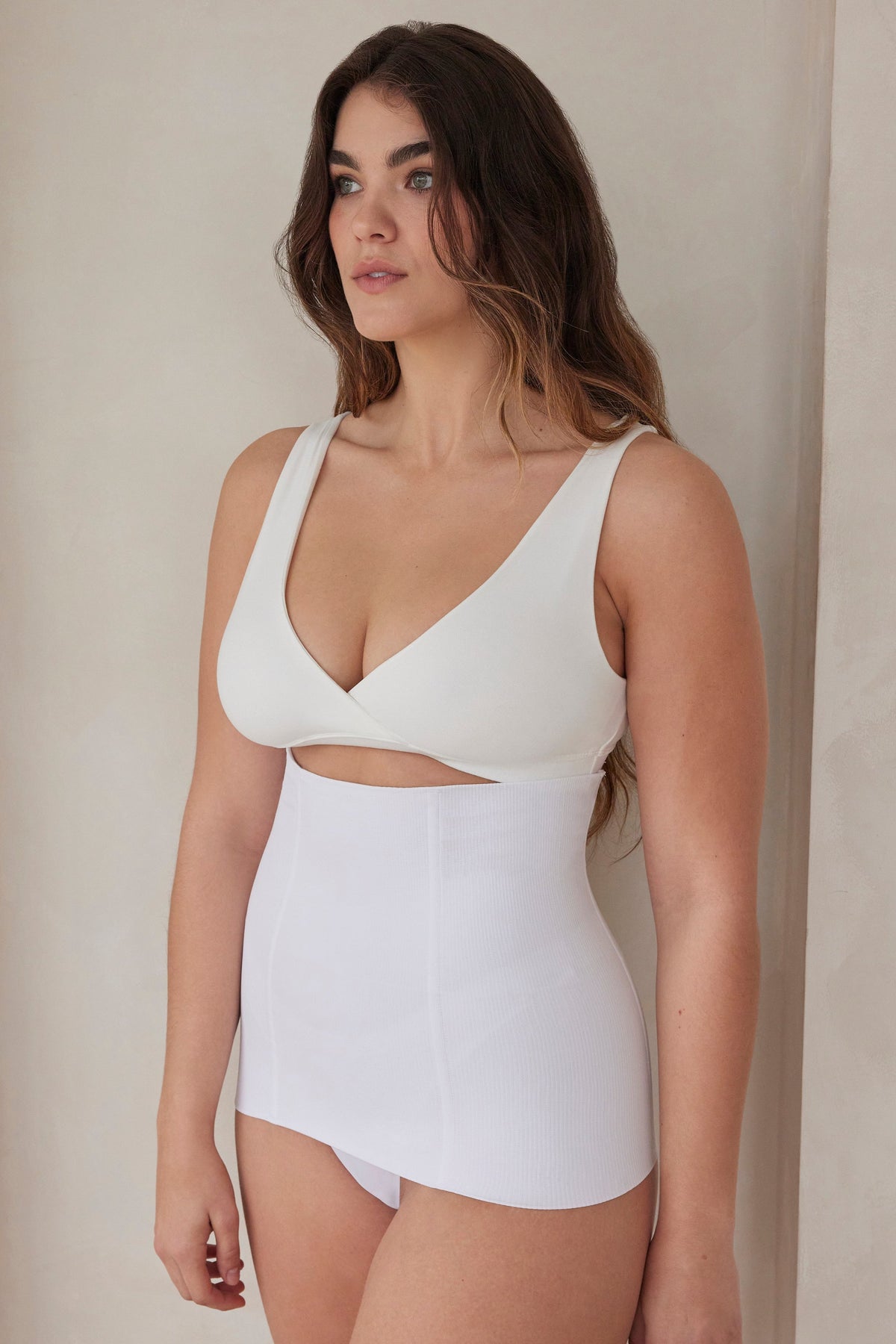Woman wearing a white reversible comfy bra and support waist trainer against a beige wall