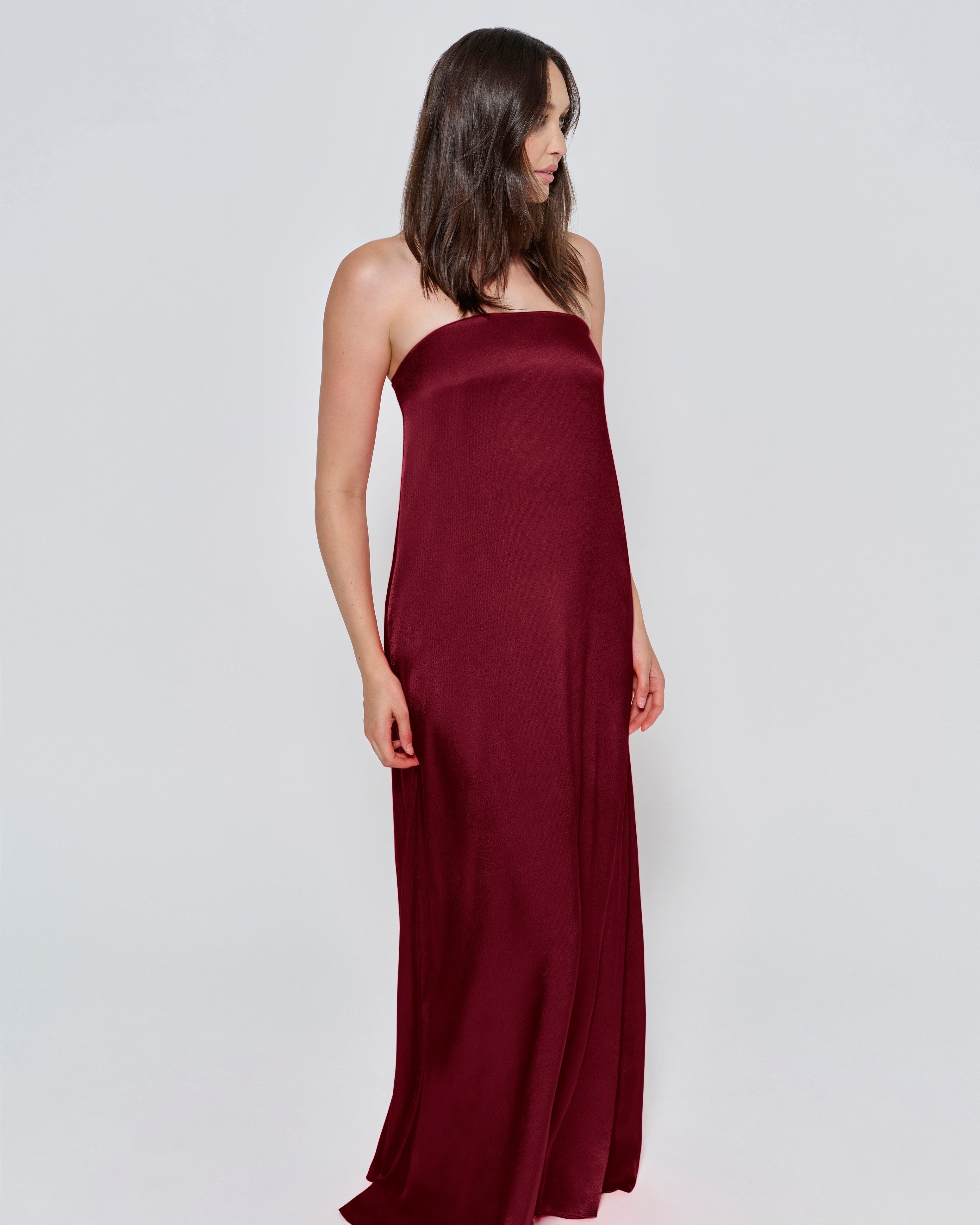 Pregnant woman wearing a Bumpsuit brushed satin gown in a Burgundy color against a white background