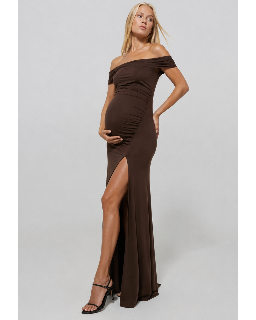 Pregnant woman wearing a chocolate coloured Bumspuit off the shoulder evening dress with a high leg split, set against a white background.
