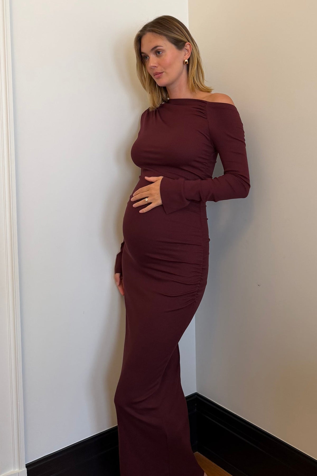 A pregnant woman wearing a long-sleeve, off-the-shoulder draped burgundy maxi maternity dress, standing in a corner of a room with off-white walls and dark baseboards.