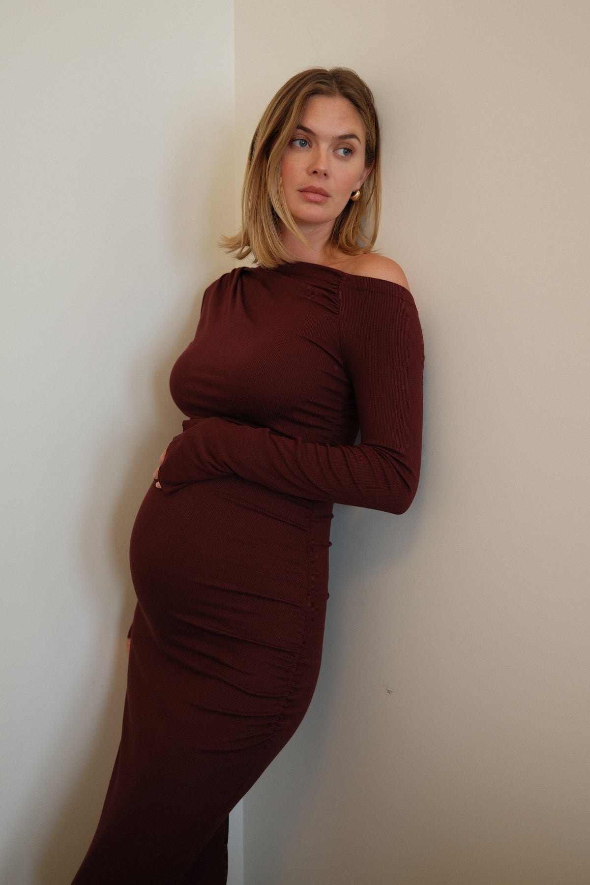 A woman with blonde hair wearing a long-sleeved, off-the-shoulder burgundy maternity dress, posing against a plain off-white wall.