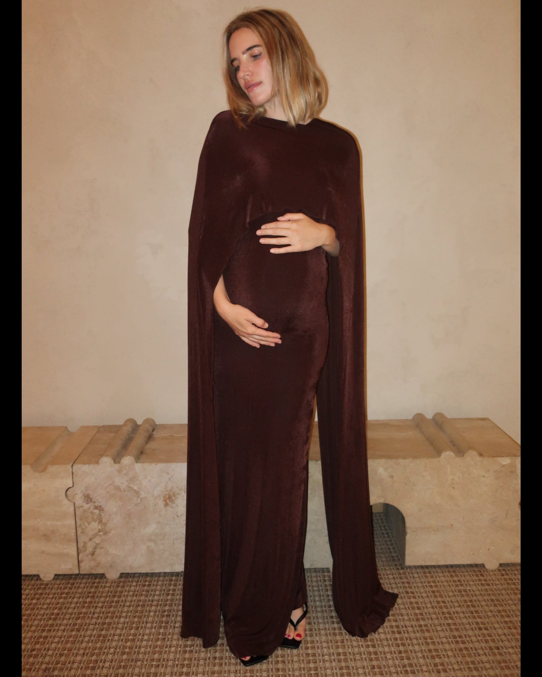 A pregnant woman wearing a Bumpsuit maxi dress with a floor length cape detail in chocolate brown against a neutral setting