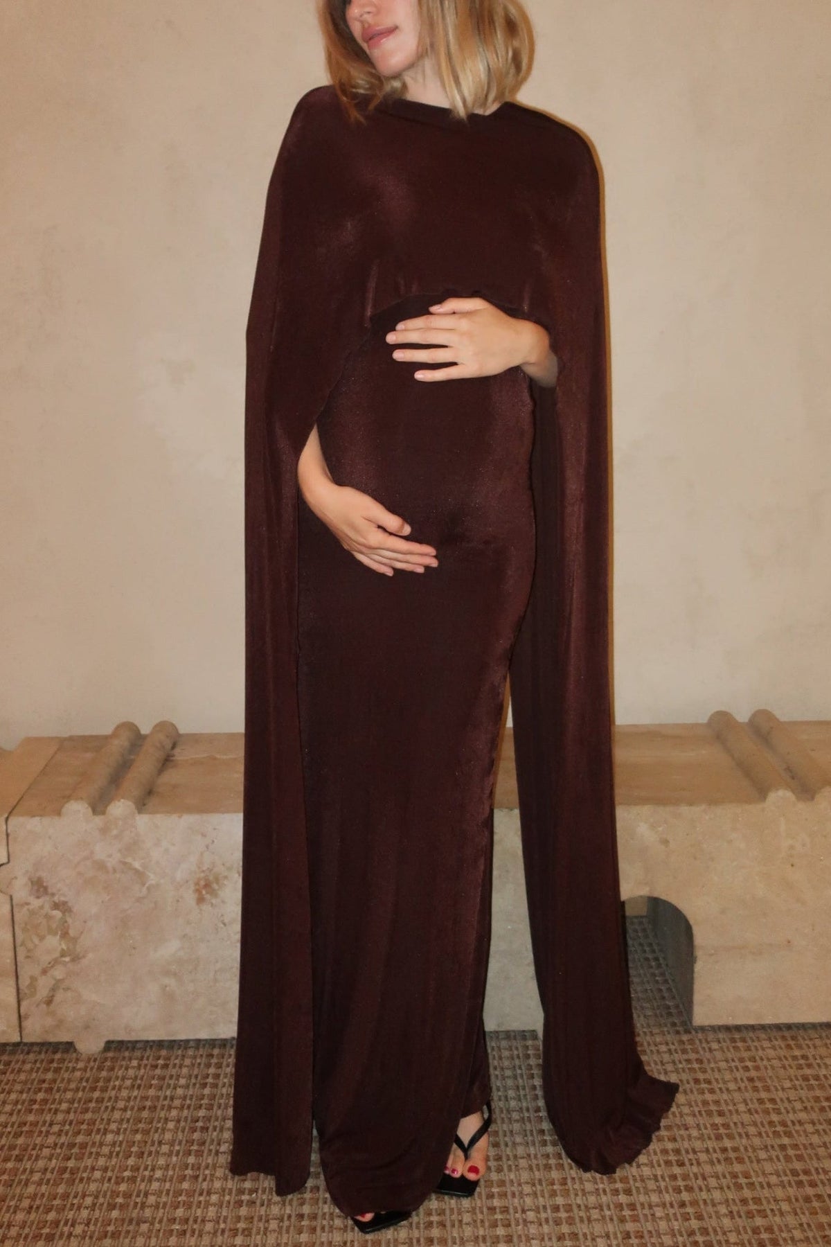 A pregnant woman wearing the Aubrey Bumpsuit maxi dress with a floor length cape detail in chocolate brown against a neutral setting