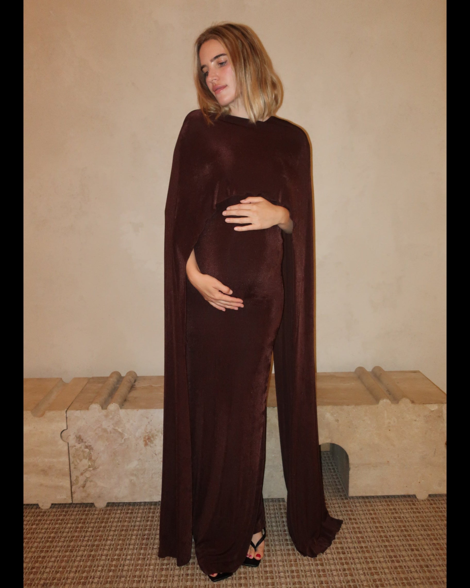 A pregnant woman wearing a Bumpsuit maxi dress with a floor length cape detail in chocolate brown against a neutral setting