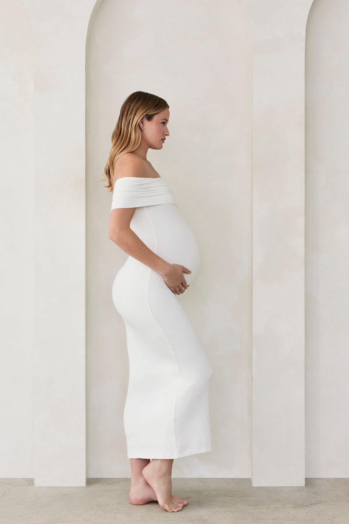 Pregnant woman wearing Bumpsuits off the shoulder Ivory Bianca maternity maxi dress standing  against a beige wall