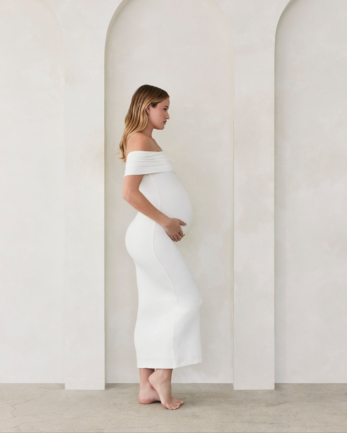 Pregnant woman wearing Bumpsuits off the shoulder Ivory Bianca maternity maxi dress standing  against a beige wall
