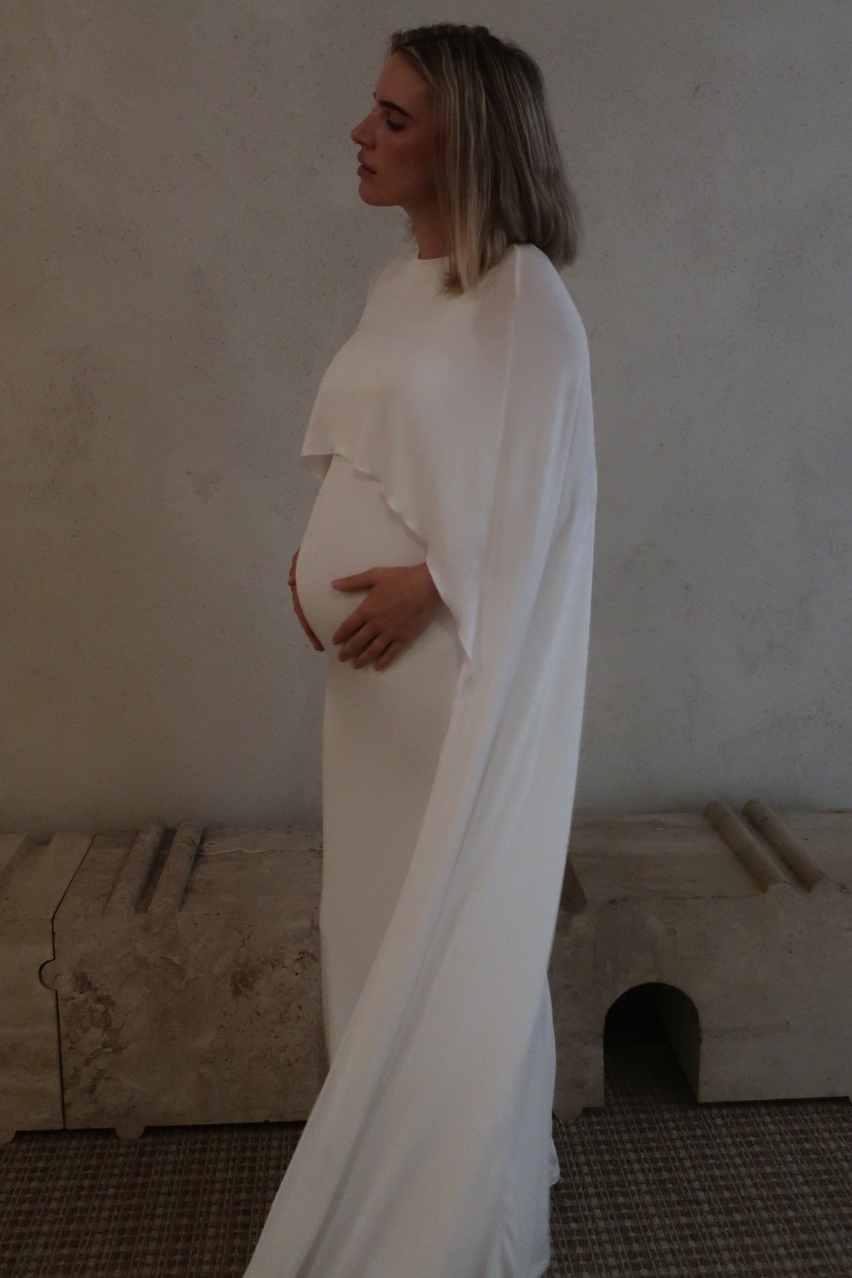 Side view of a pregnant woman wearing a maxi cape style dress in Ivory  against a beige background 