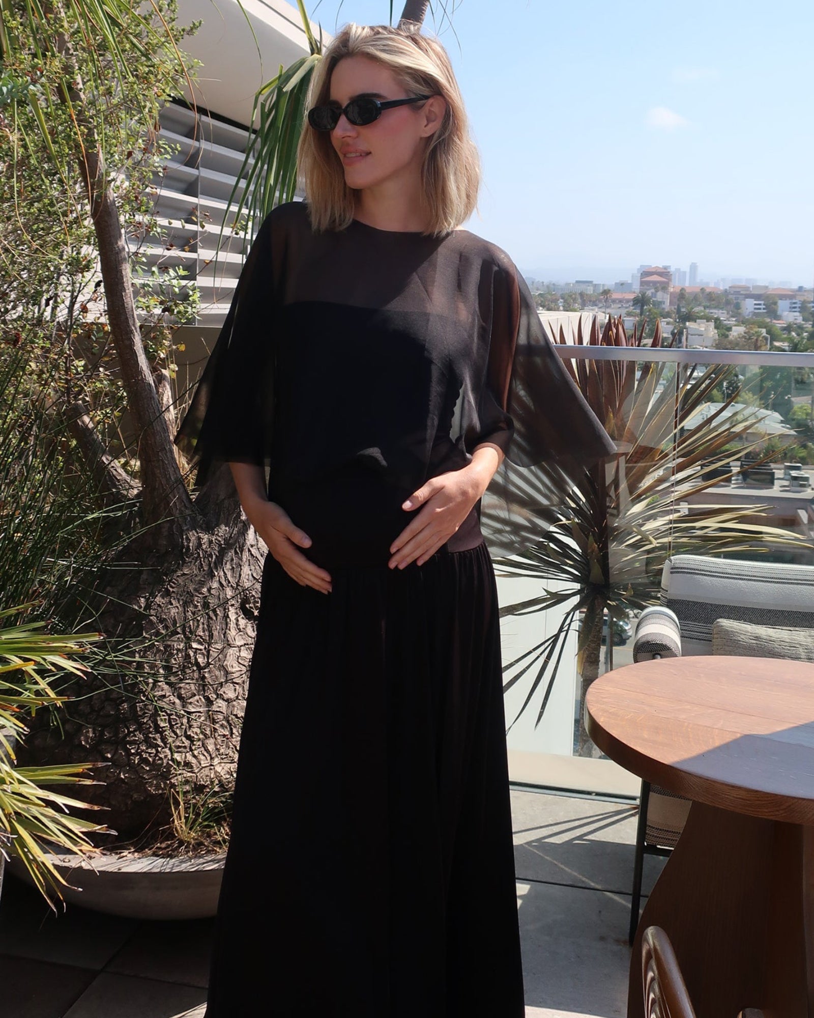 A pregnant woman wearing Bumpsuits Maria maternity maxi dress with a chiffon capulet and sunglasses, standing on a sunny rooftop terrace with plants and a city view in the background.