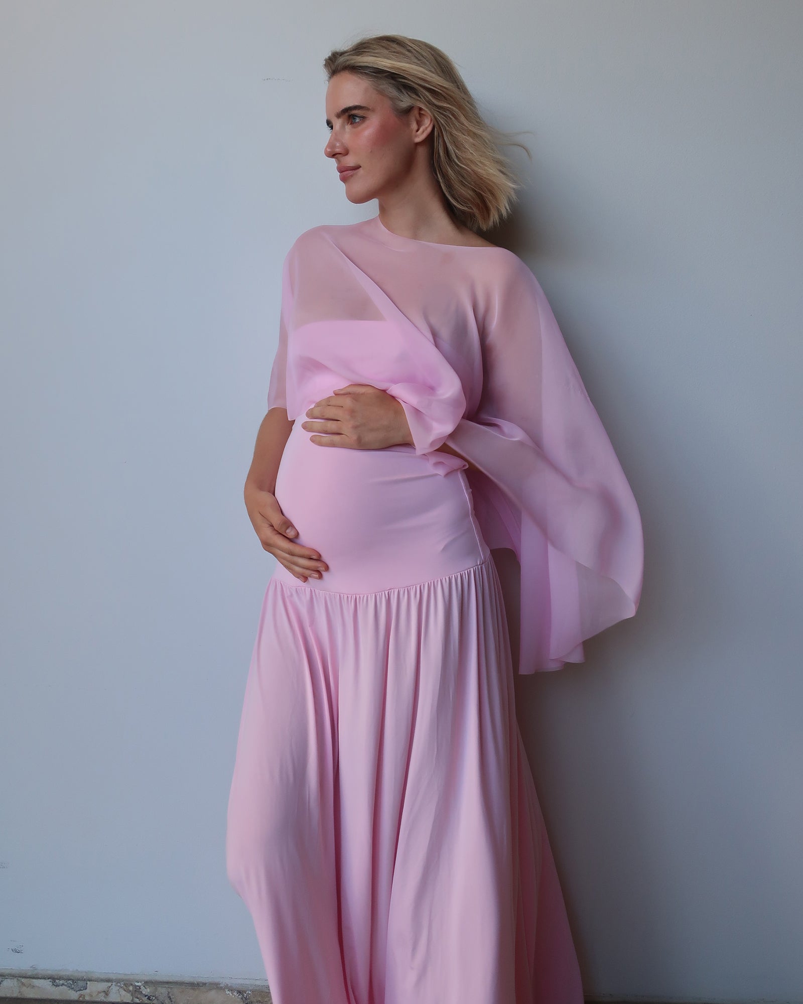 A pregnant woman wearing a light pink maternity dress with a draped  chiffon cape, standing against a plain light gray wall.