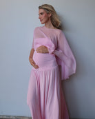 A pregnant woman wearing a light pink maternity dress with a draped  chiffon cape, standing against a plain light gray wall.