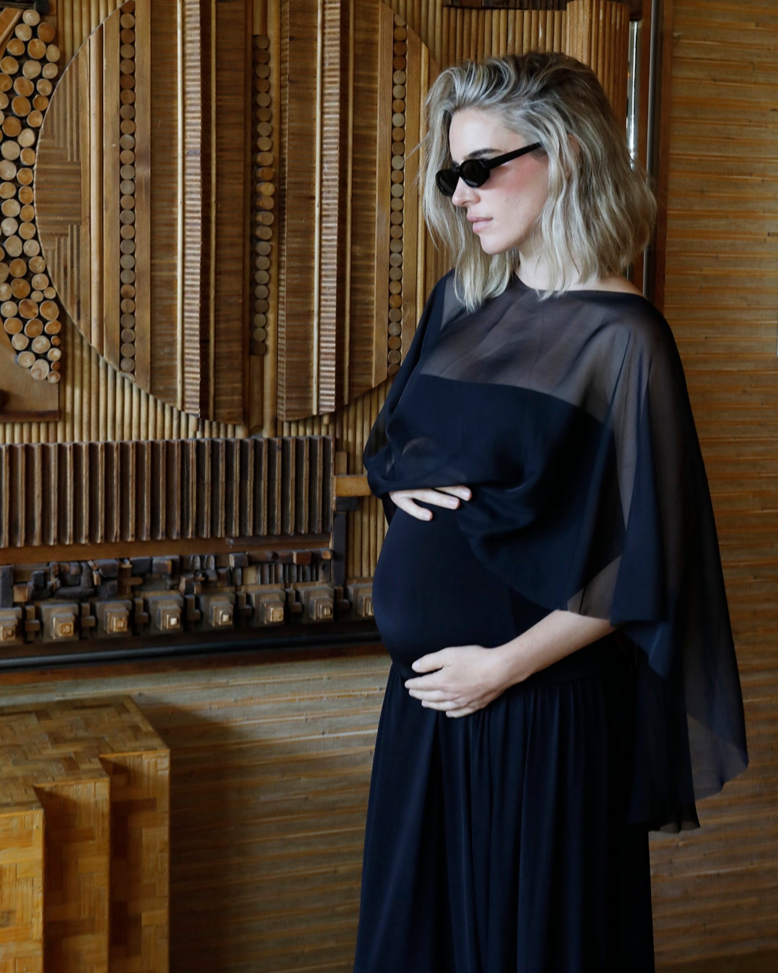 A pregnant woman wearing a black maternity gown with chiffon capelet by Bumpsuit and black sunglasses, standing in front of a wooden geometric wall art piece.