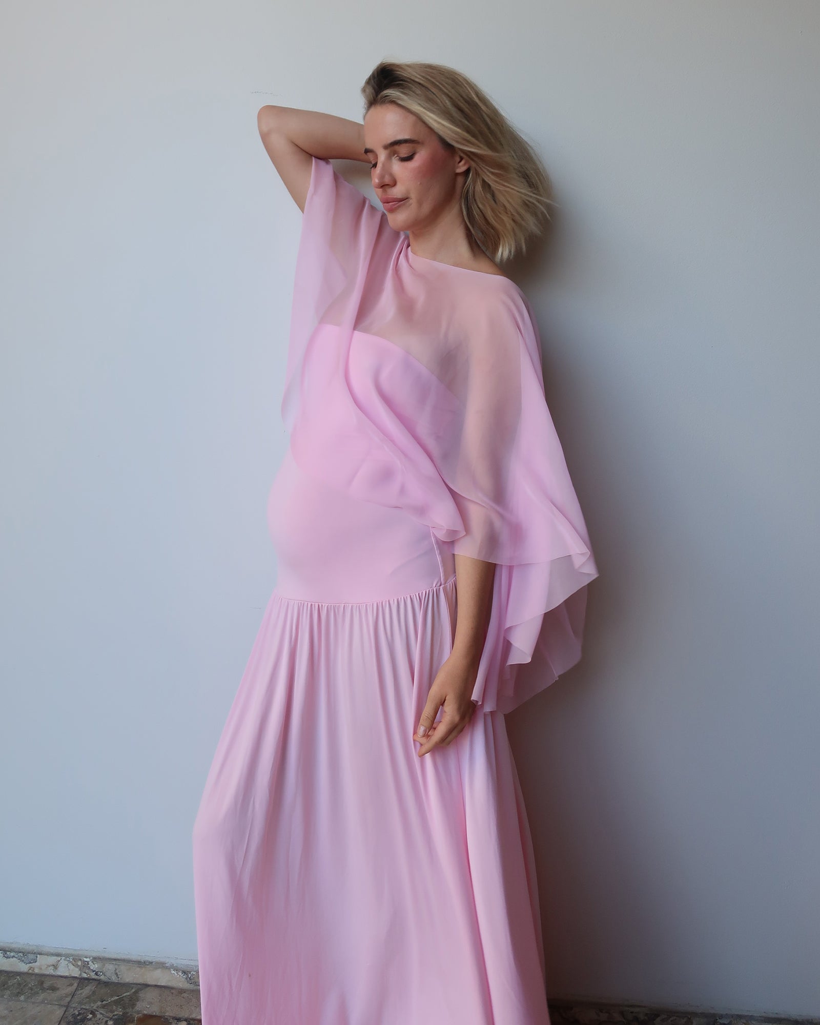 A pregnant woman wearing a Bumpsuit pink maternity dress with a sheer draped cape, posing against a plain light gray wall.
