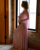 A pregnant woman in a Bumpsuit pink sheer maxi dress with a chiffon cape in profile by a window in a room with floral wallpaper and wood trim.