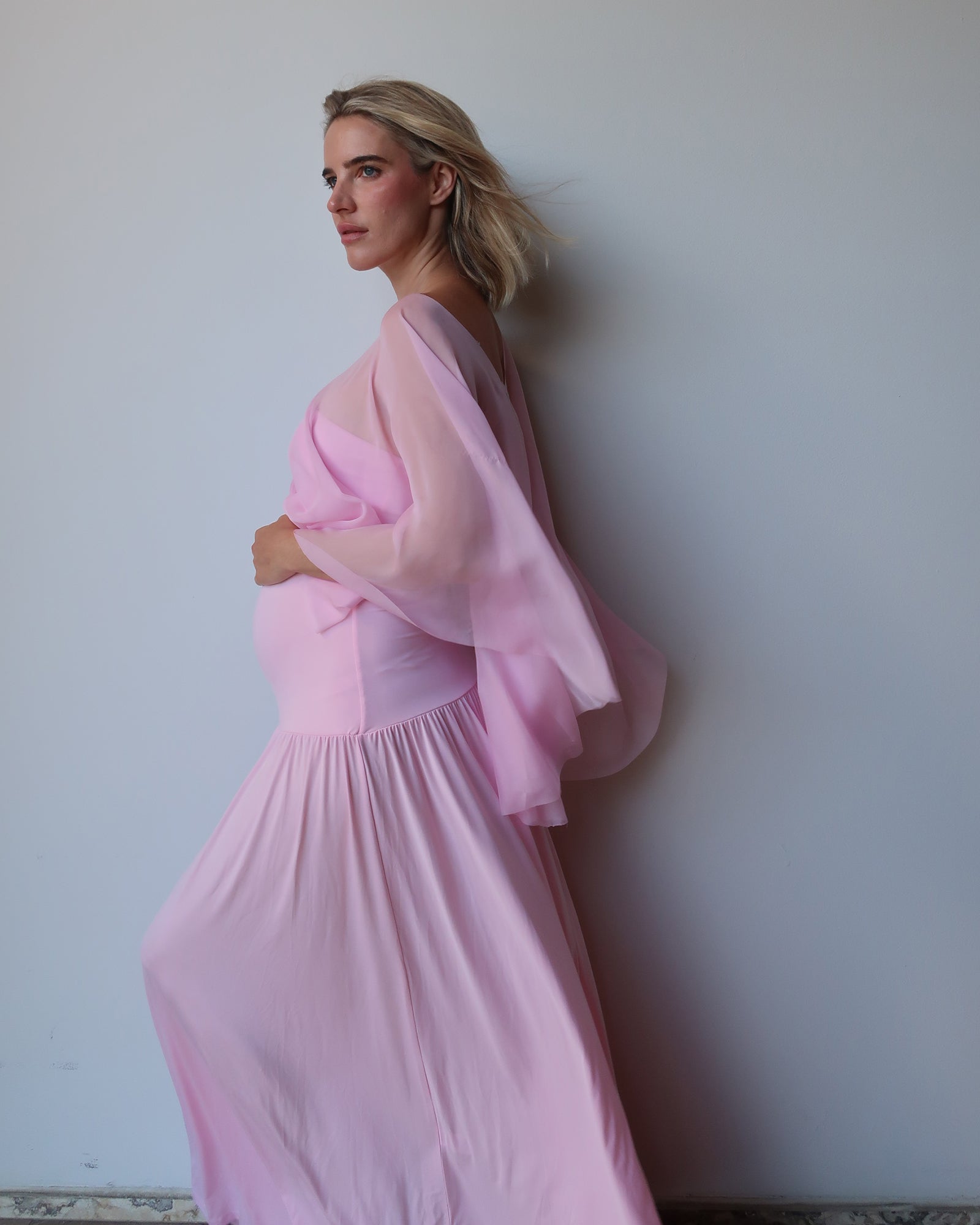 A pregnant woman wearing a flowing light pink maternity dress with chiffon cape by Bumpsuit, standing against a plain light gray wall.