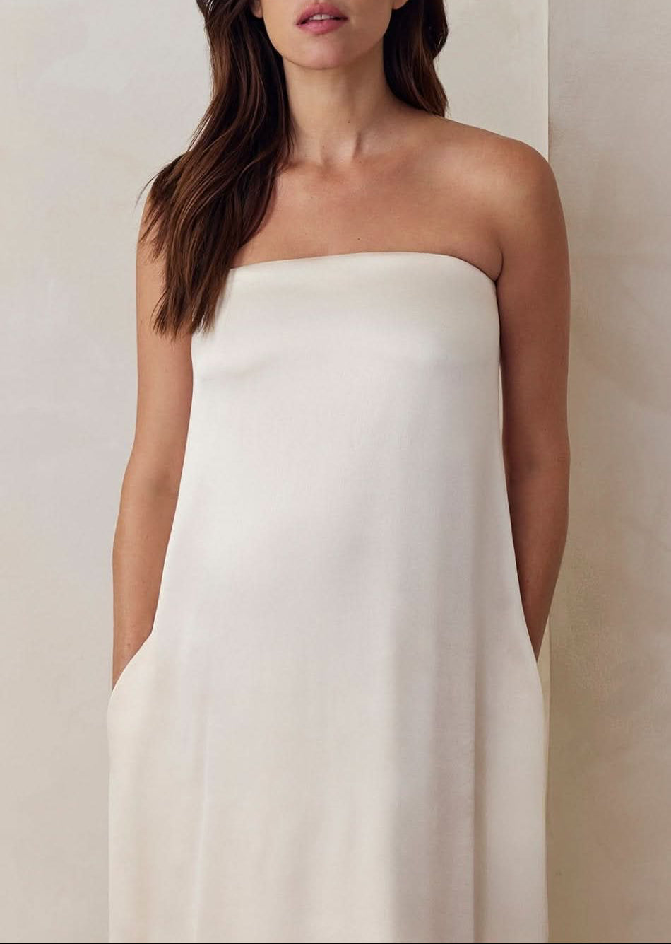 A Pregnant model wearing the Bumpsuit brushed satin gown in ivory against a beige feature wall.