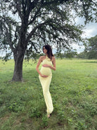Pregnant woman in a light yellow dress standing in a grassy field with trees.