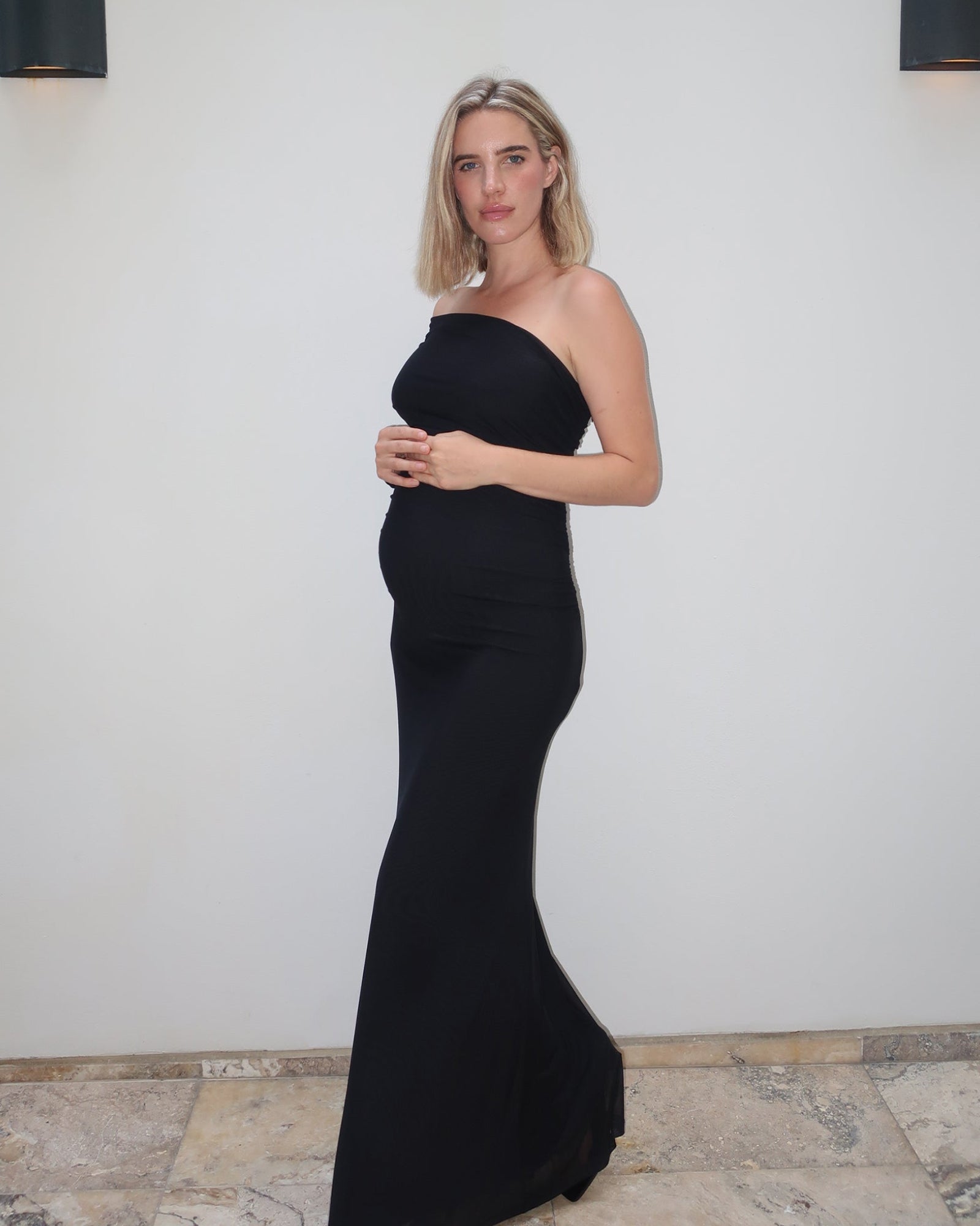 A pregnant woman wearing  the Vanessa Bumpsuit off the shoulder gown, standing against a white wall with stone tile flooring.