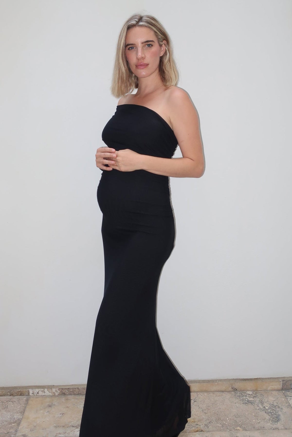 A pregnant woman wearing  the Vanessa Bumpsuit off the shoulder gown, standing against a white wall with stone tile flooring.