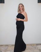 A pregnant woman wearing  the Vanessa Bumpsuit off the shoulder gown, standing against a white wall with stone tile flooring.