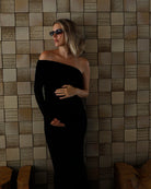A pregnant woman wearing a Bumpsuit black off-the-shoulder floor-length gown and dark sunglasses stands against a beige tiled wall with wooden benches at the bottom.