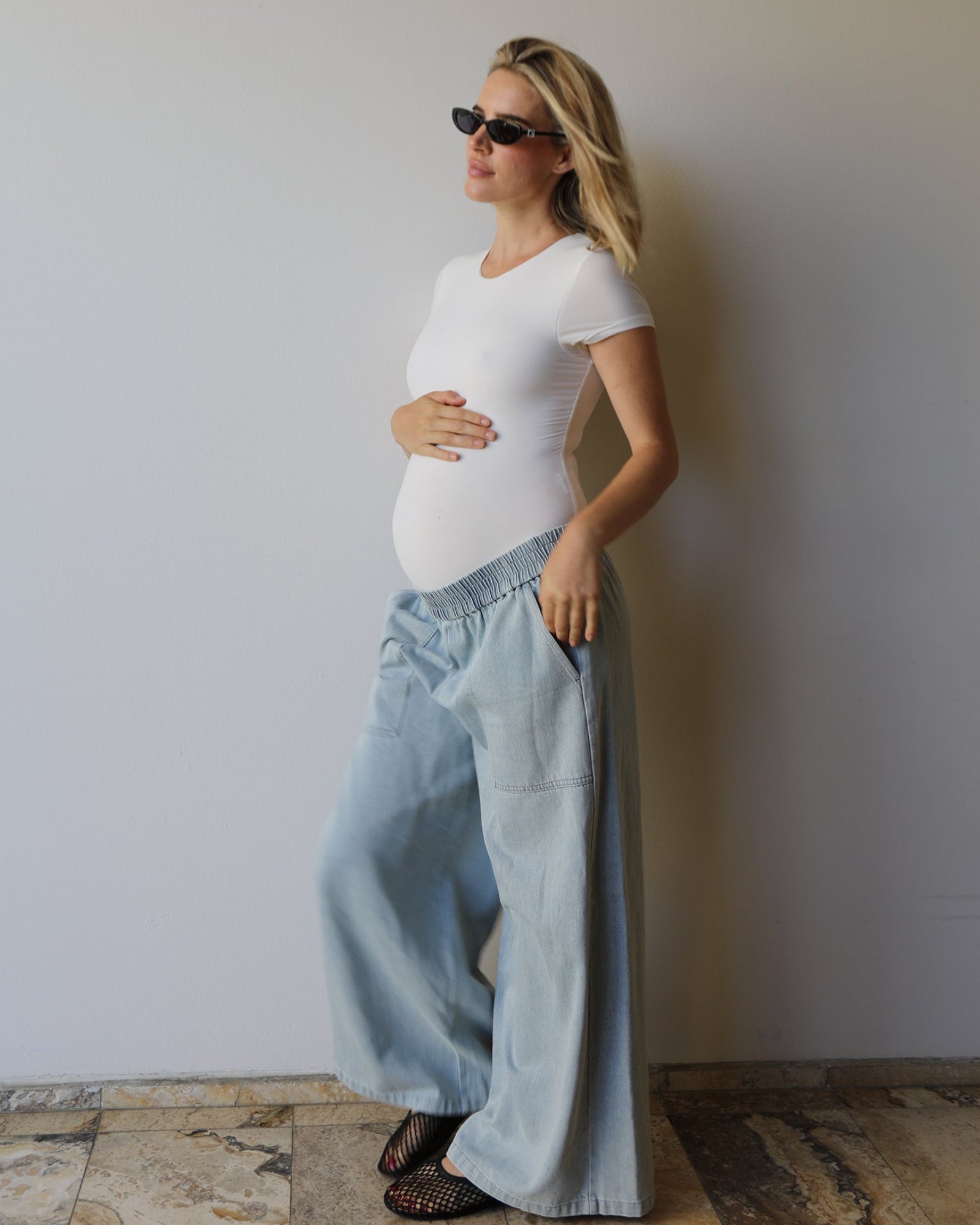 A pregnant woman wearing a Bumpsuit white short-sleeve everyday shirt and light blue wide-leg denim pants, standing against a plain light gray wall on a stone-tiled floor.