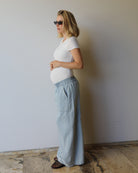 A pregnant woman in side profile wearing the Bumpsuit Everyday shirt and light blue wide-leg denim pants, standing against a plain light gray wall.