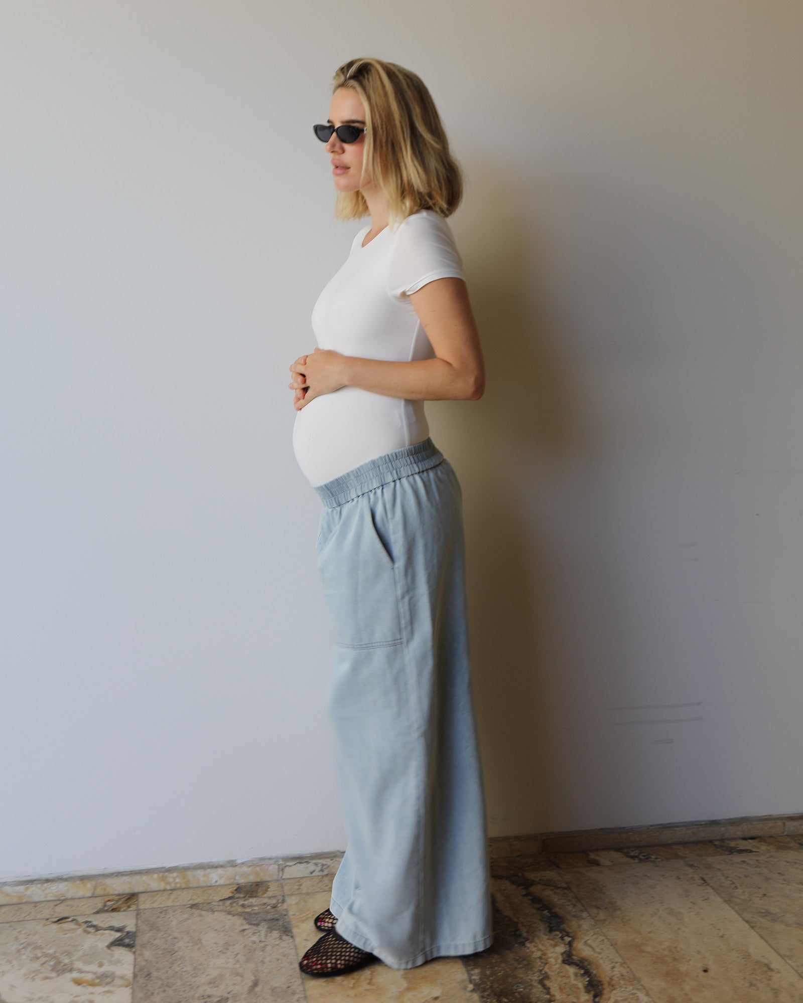 A pregnant woman in side profile wearing the Bumpsuit Everyday shirt and light blue wide-leg denim pants, standing against a plain light gray wall.