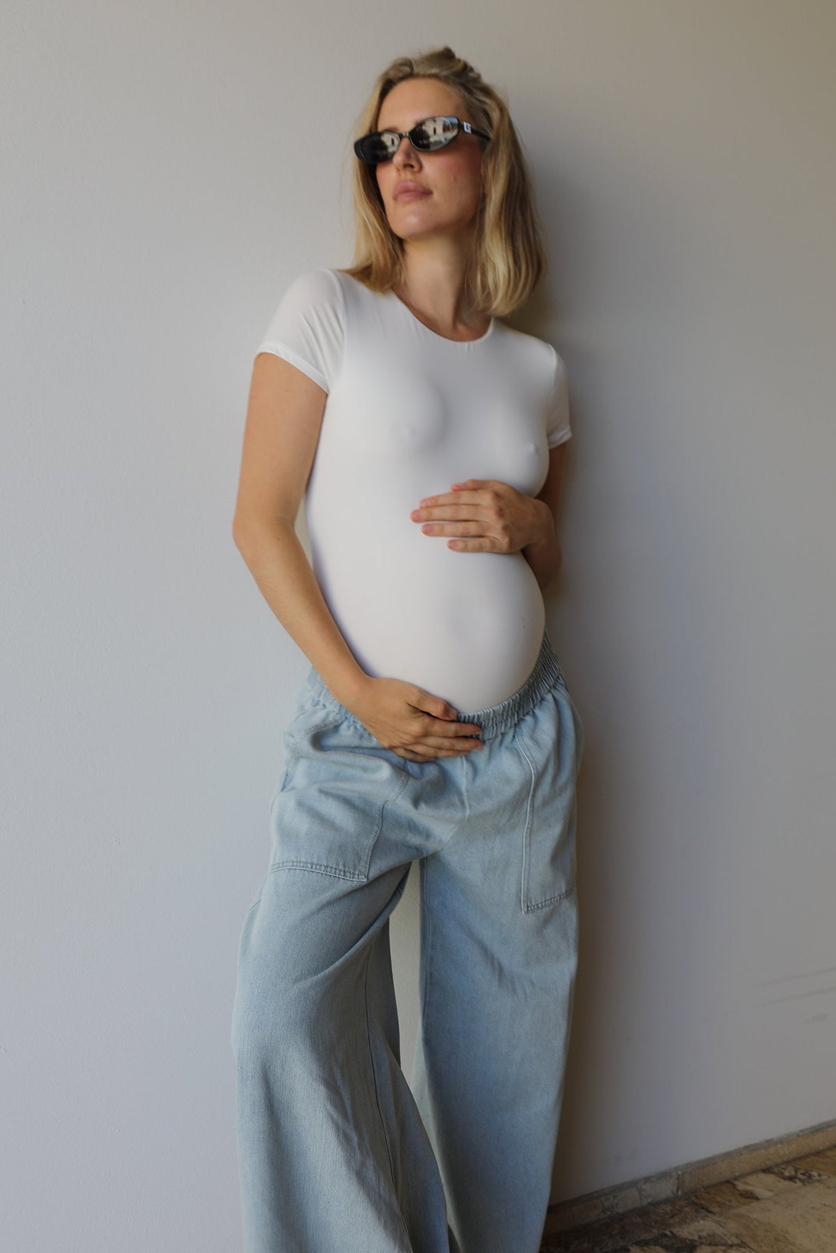 A pregnant woman wearing a fitted white short-sleeve t-shirt and light blue wide-leg jeans, standing against a plain light gray wall.