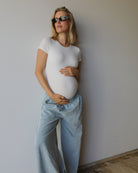 A pregnant woman wearing a fitted white short-sleeve t-shirt and light blue wide-leg jeans, standing against a plain light gray wall.
