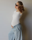 A pregnant woman wearing a white short-sleeve bodysuit and light blue wide-leg jeans by Bumpsuit, posing in profile against a plain light gray wall.