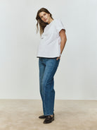 Pregnant woman wearing a white top and blue jeans standing against a plain background