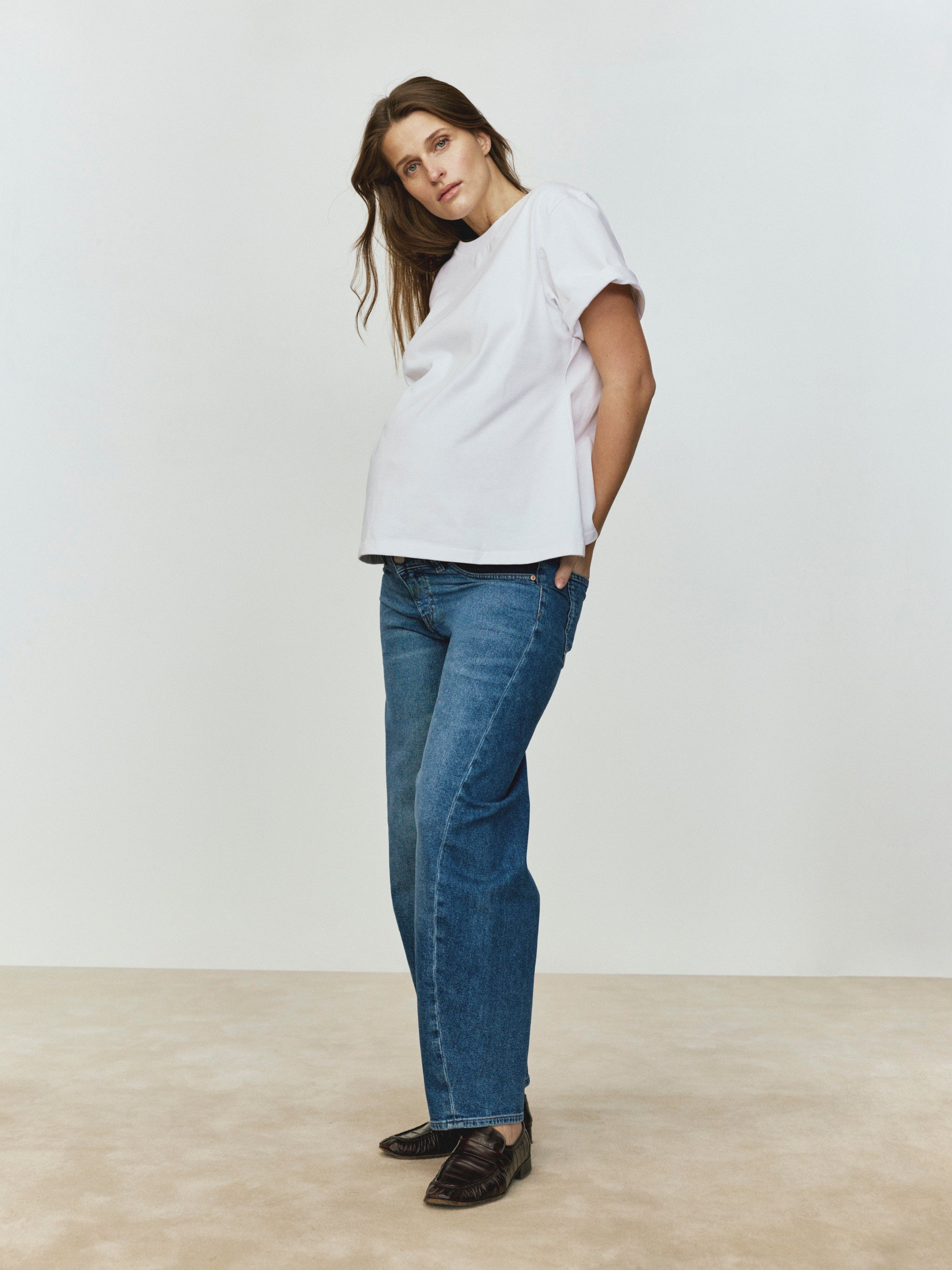 Pregnant woman wearing a white top and blue jeans standing against a plain background