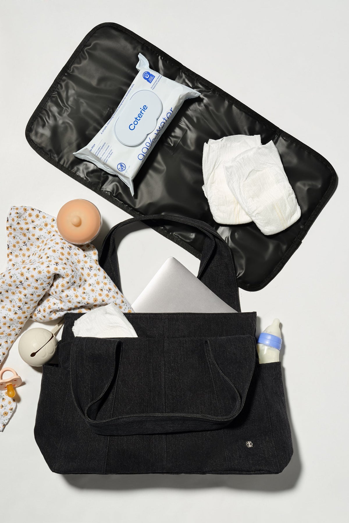 A black diaper bag shown open in a flat-lay arrangement, filled with baby essentials including a patterned baby shirt, wipes, diapers, a pacifier, and a bottle, on a light gray background.