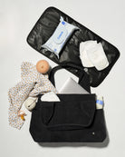 A black diaper bag shown open in a flat-lay arrangement, filled with baby essentials including a patterned baby shirt, wipes, diapers, a pacifier, and a bottle, on a light gray background.