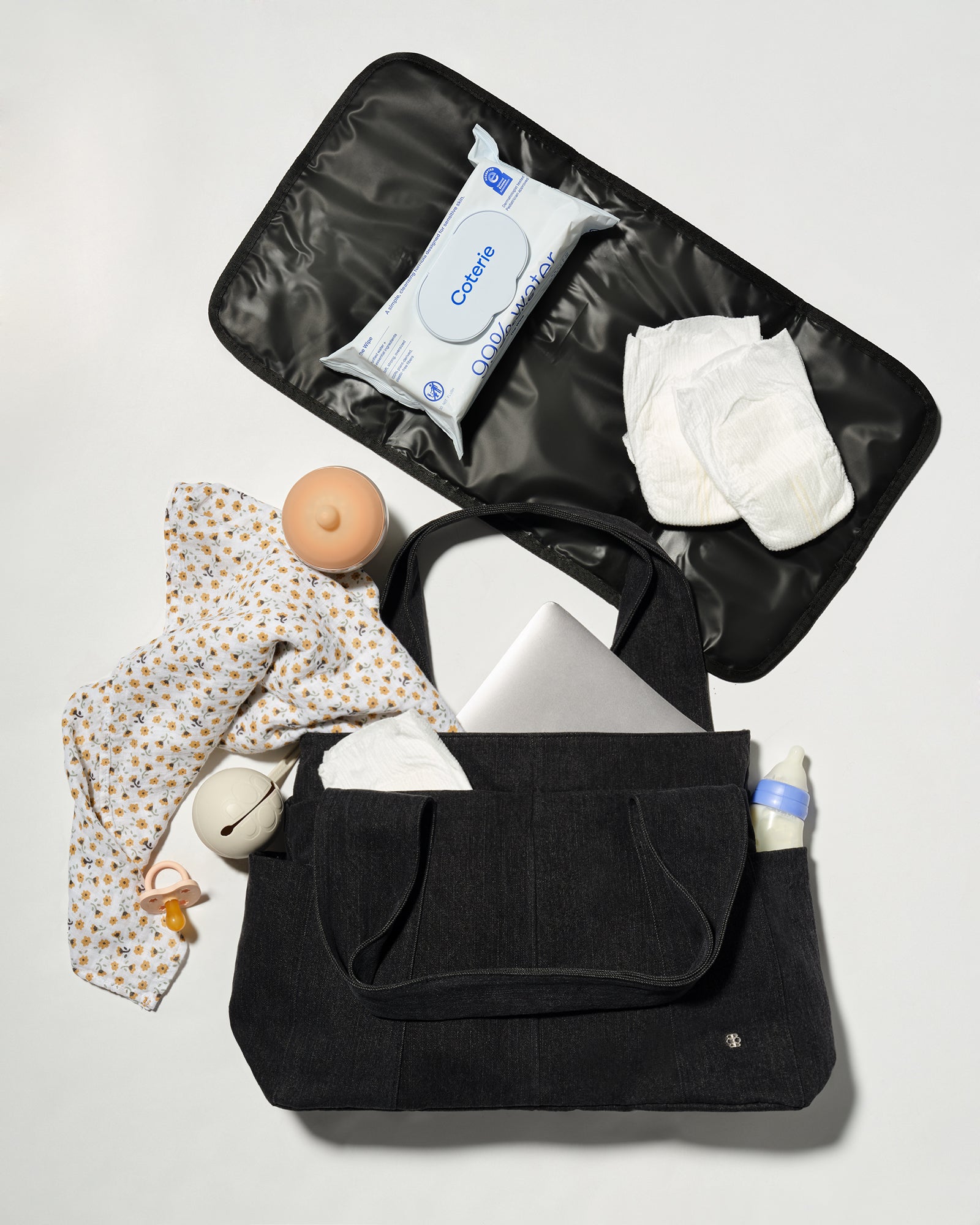 A black diaper bag shown open in a flat-lay arrangement, filled with baby essentials including a patterned baby shirt, wipes, diapers, a pacifier, and a bottle, on a light gray background.