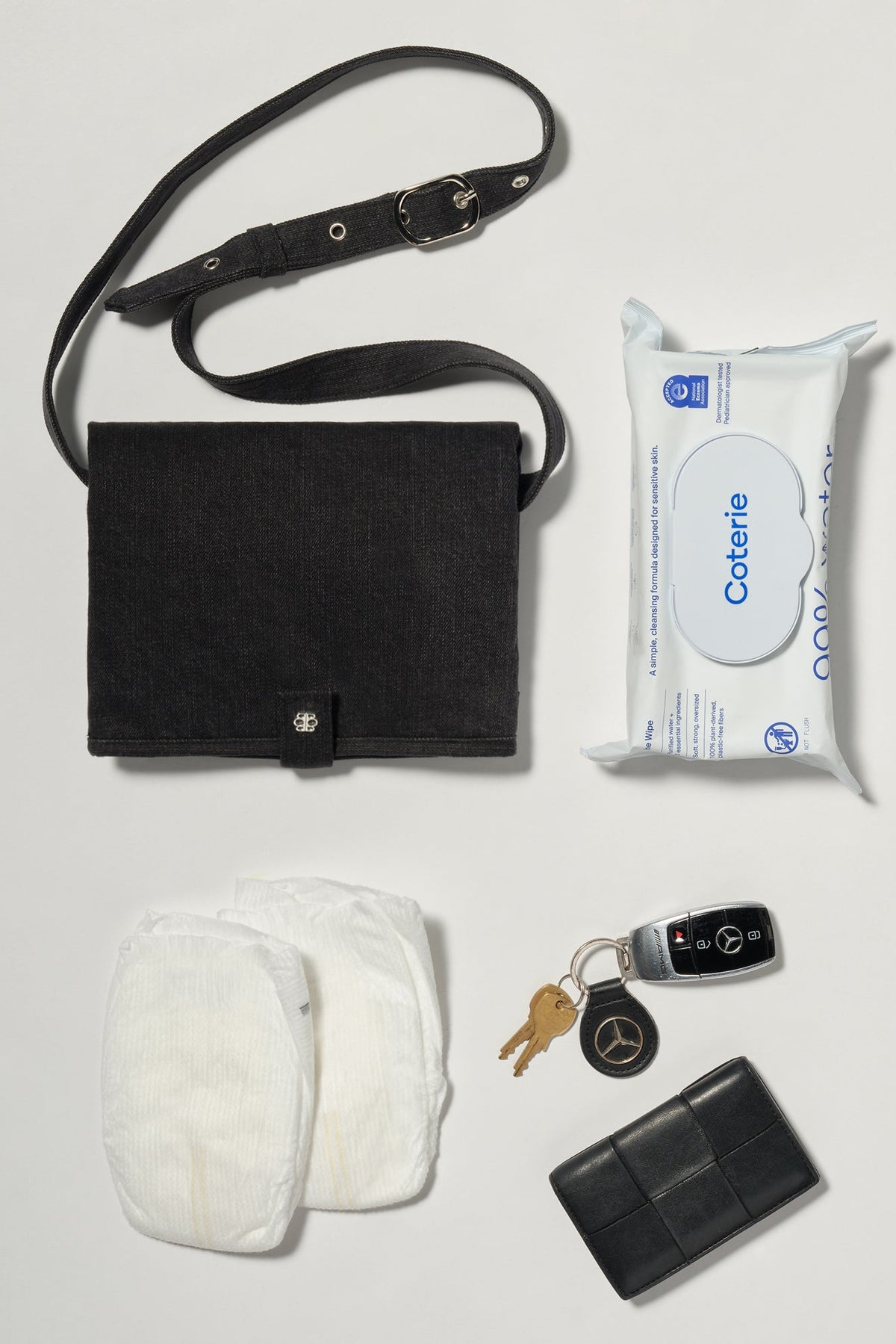 Flat lay of the Mom-On-The-Go Bag showcasing black denim fabric and adjustable strap beside wipes, diapers, keys, and wallet