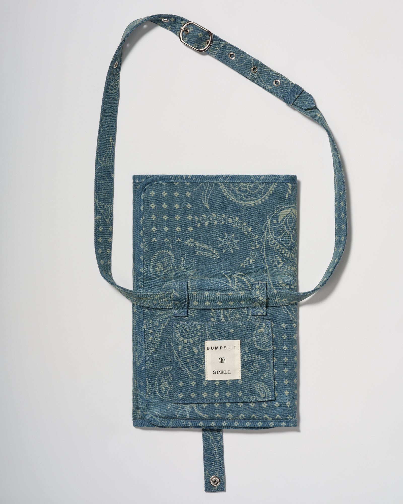 A blue denim crossbody diaper change mat bag by with a paisley and geometric pattern by Spell and Bumpsuit,, shown on a light gray background.