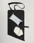 A black fabric diaper bag or organizer pouch with a top strap, shown on a light gray background. The pouch has a front pocket holding a package of baby wipes and a white diaper hanging below.