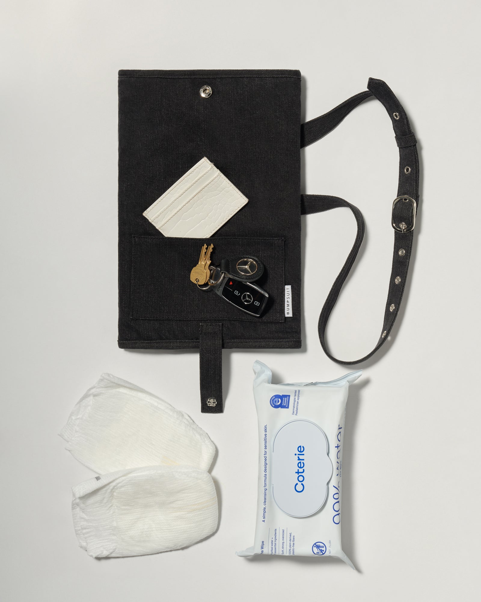 A black denim roll up diaper change mat bag by Spell and Bumpsuit, shown on a light gray background with wipes, diapers, keys, and wallet