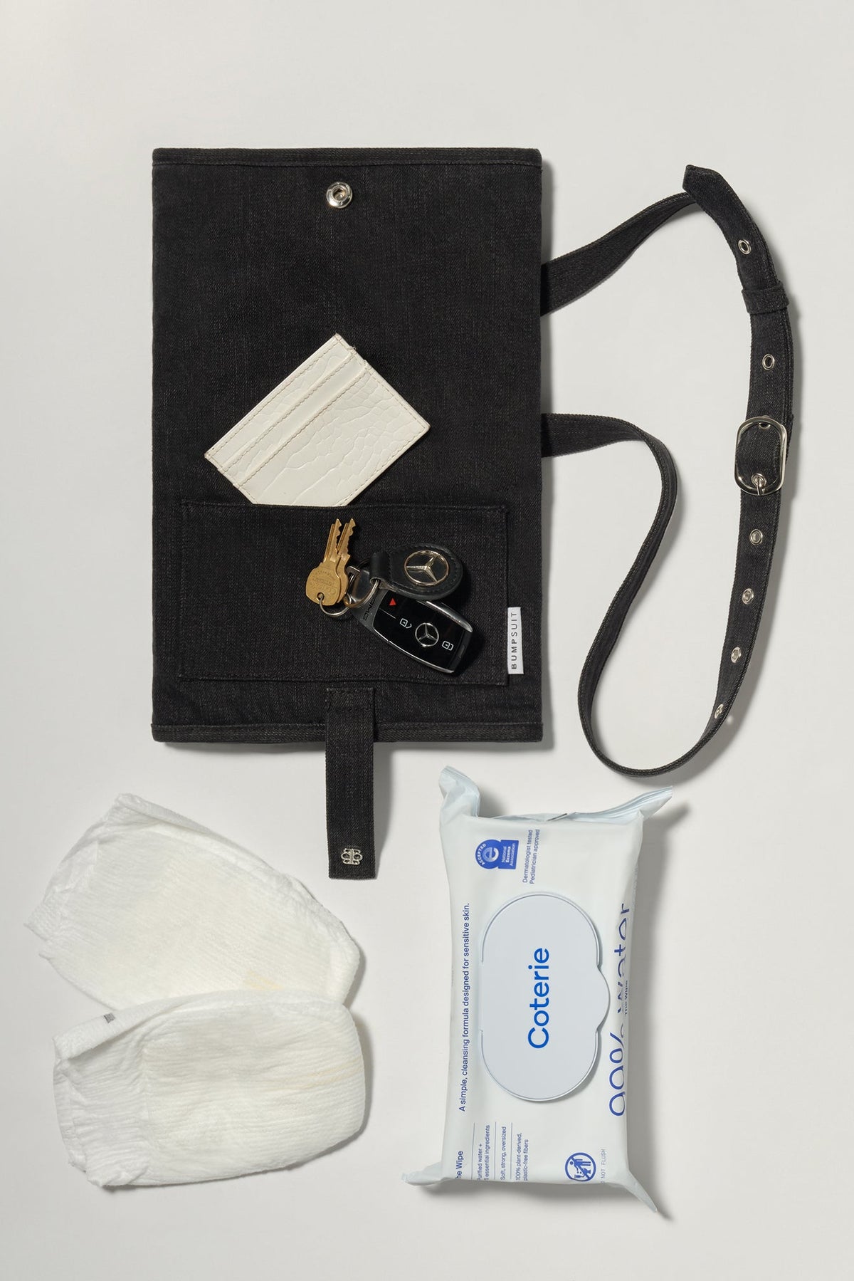 A black denim roll up diaper change mat bag by Spell and Bumpsuit, shown on a light gray background with wipes, diapers, keys, and wallet