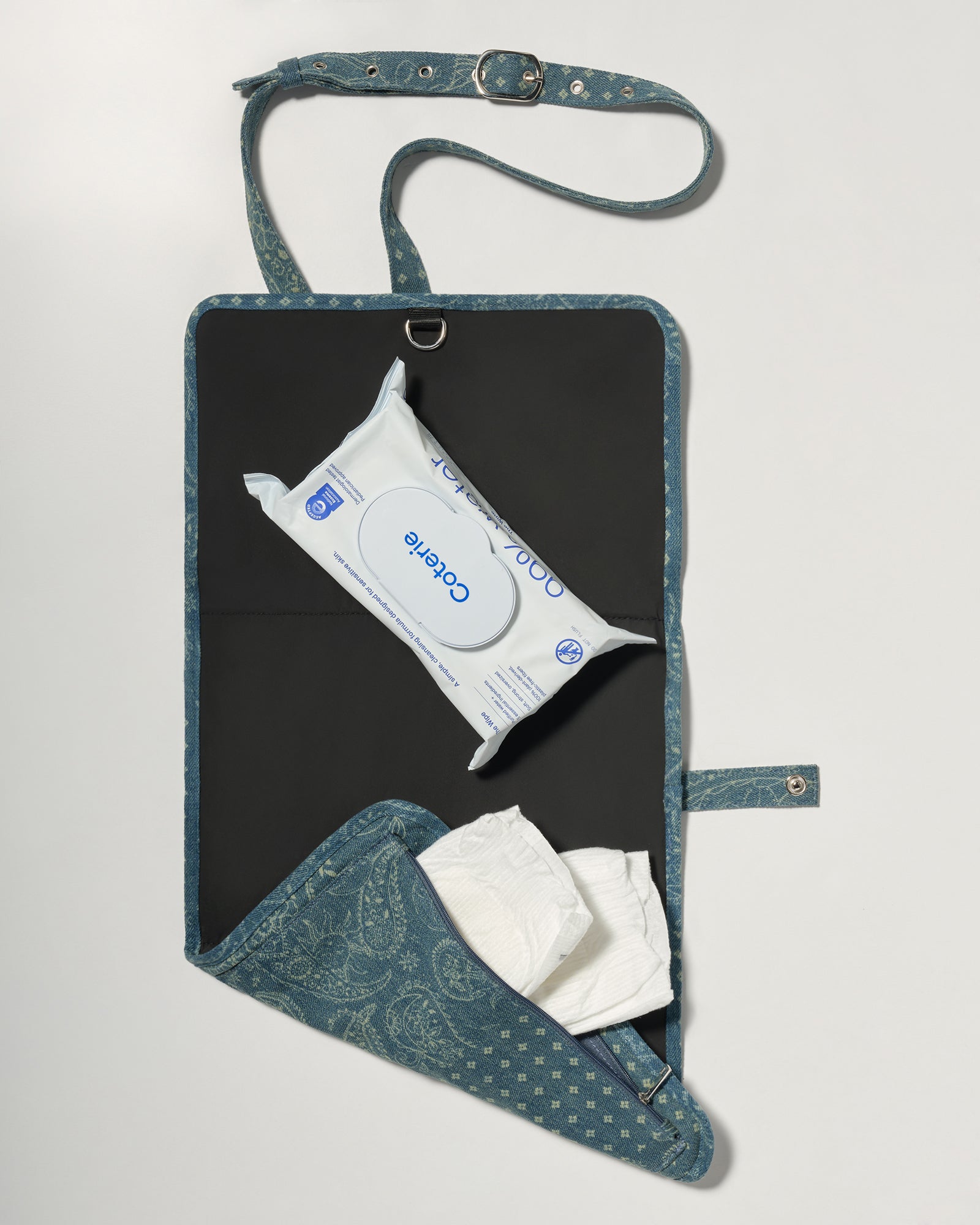 A blue patterned diaper wrap bag with a black interior, shown open on a light gray background. It contains a pack of wipes and white diapers.