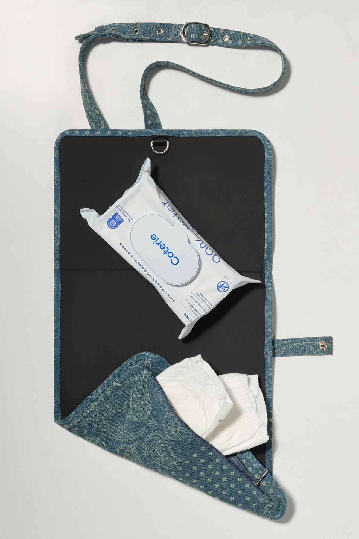 A blue patterned diaper wrap bag with a black interior, shown open on a light gray background. It contains a pack of wipes and white diapers.