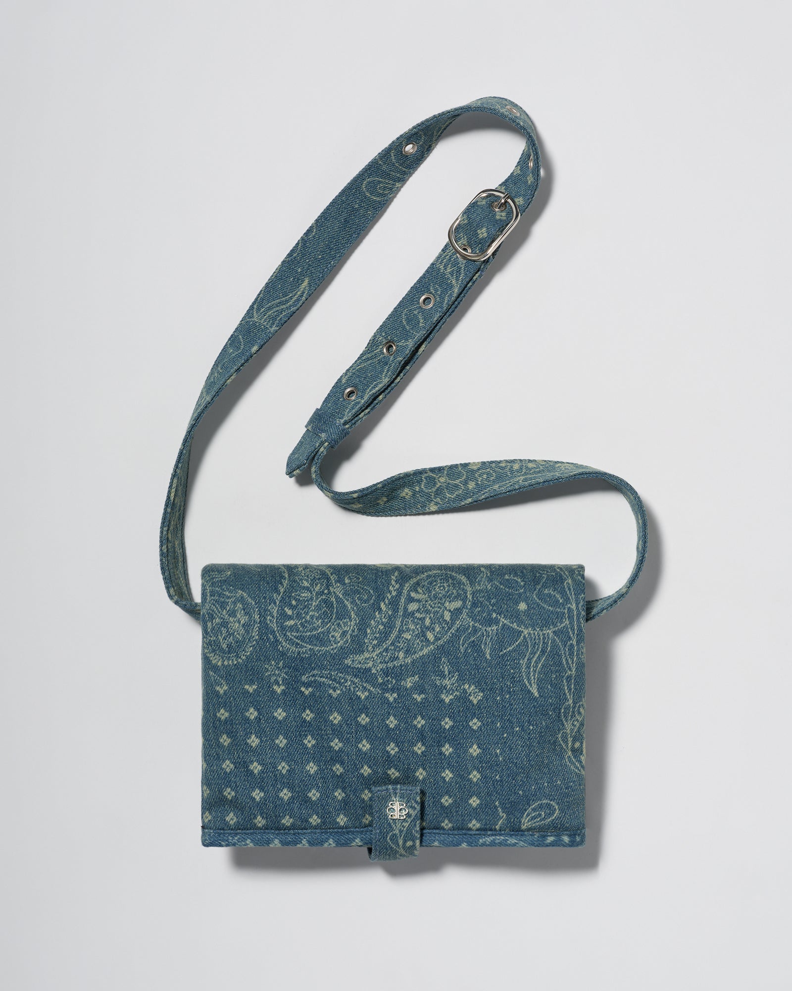 A blue denim crossbody diaper change mat bag with a paisley and geometric pattern by Spell and Bumpsuit,, shown on a light gray background.