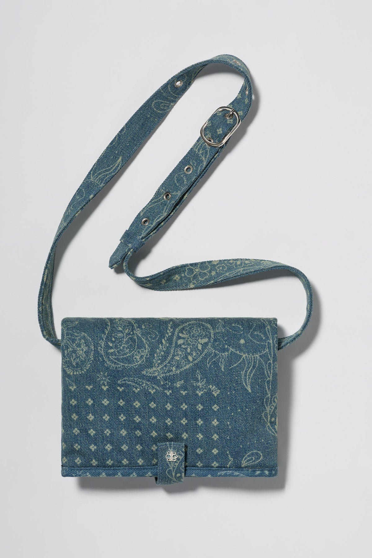 A blue denim crossbody diaper change mat bag with a paisley and geometric pattern by Spell and Bumpsuit,, shown on a light gray background.