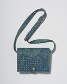 A blue denim crossbody diaper change mat bag with a paisley and geometric pattern by Spell and Bumpsuit,, shown on a light gray background.
